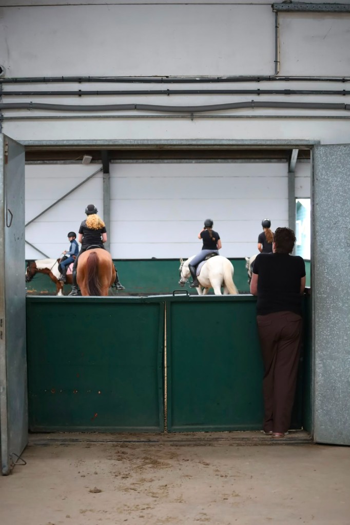 Instructeur kijkt vanaf de poort naar ruiters tijdens een les in de binnenbak